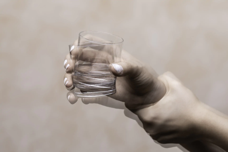 A hand shaking while holding a glass of water. The hand is clearly affected by Parkinson's Disease, as the tremors are visible.